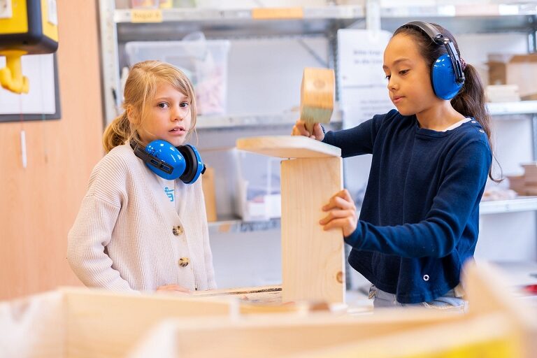 Zwei Kinder in der Junior Uni, beide mit Hörschutz, das eine hämmert konzentriert Holzbretter zusammen, das andere schaut in die Kamera.