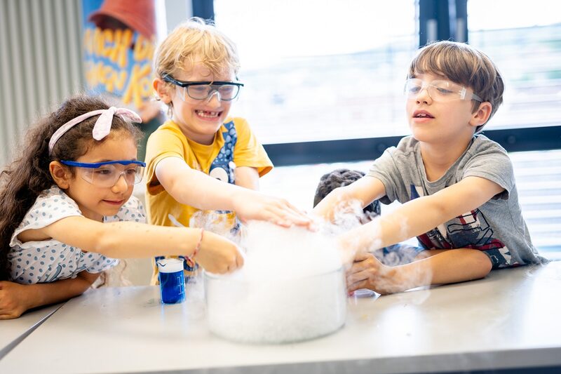 Drei Kinder halten ihre Hände in wabernden Dampf, der aus einem Glas auf dem Tisch steigt. Die Kinder haben Schutzbrillen auf und lachen.