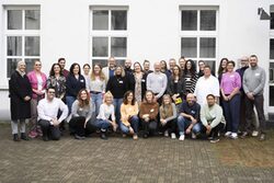 Gruppenfoto von etwa 30 Personen vor einem weißen Gebäude mit Fenstern. Die Personen stehen und knien in mehreren Reihen, einige lächeln, andere schauen neutral in die Kamera. Sie tragen unterschiedliche, meist lässige Kleidung. Es handelt sich um eine diverse Gruppe von Männern und Frauen in verschiedenen Altersgruppen und Stilen.