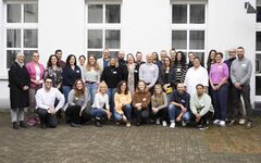 Gruppenfoto von etwa 30 Personen vor einem weißen Gebäude mit Fenstern. Die Personen stehen und knien in mehreren Reihen, einige lächeln, andere schauen neutral in die Kamera. Sie tragen unterschiedliche, meist lässige Kleidung. Es handelt sich um eine diverse Gruppe von Männern und Frauen in verschiedenen Altersgruppen und Stilen.