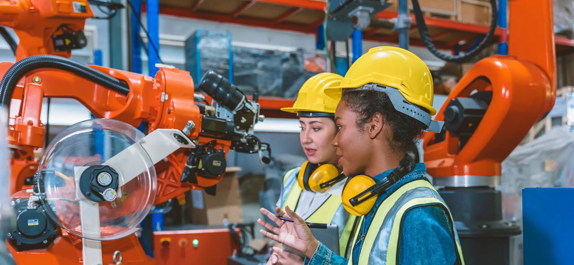 Zwei Ingenieurinnen mit Schutzhelmen und Sicherheitswesten arbeiten an einem Industrieroboter in einer Fabrikumgebung. Eine der Frauen hält ein Tablet, während sie den Roboter inspizieren und besprechen.