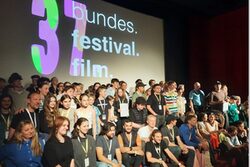 Eine große Gruppe von Menschen, darunter die Teilnehmer des Medienprojekts Wuppertal, posiert für ein Foto auf der Bühne des Filmforum Duisburg beim Bundes.Festival.Film. Im Hintergrund ist das Logo des Festivals zu sehen