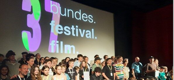 Eine große Gruppe von Menschen, darunter die Teilnehmer des Medienprojekts Wuppertal, posiert für ein Foto auf der Bühne des Filmforum Duisburg beim Bundes.Festival.Film. Im Hintergrund ist das Logo des Festivals zu sehen.