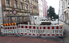 Blick auf eine Straßensperrung mit rot-weißen Bauzäunen, im Hintergrund Gründerzeithäuser, links eine baufällige Fassade mit Gerüst