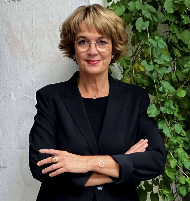 Portrait Annette Berg im schwarzen Blazer vor weißer Wand und grünem Laub