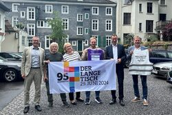 Die Organisatoren, unter anderem OB Uwe Schneidewind, halten gemeinsam ein Plakat in den Händen, mit der Aufschrift: "Der Lange Tisch, 29. Juni 2024"