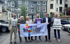 Die Organisatoren, unter anderem OB Uwe Schneidewind, halten gemeinsam ein Plakat in den Händen, mit der Aufschrift: "Der Lange Tisch, 29. Juni 2024"
