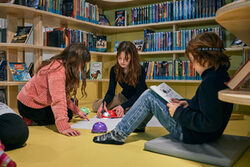 3 Mädchen sitzen auf dem Boden umringt von Bücherregalen
