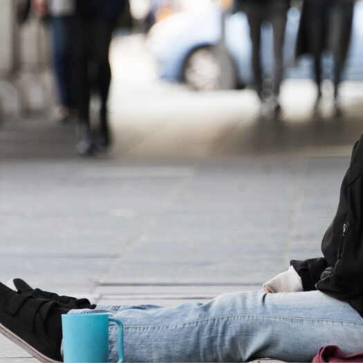 Symbolbild: Ein Mensch sitzt auf der Straße, neben ihm steht eine blaue Tasse.